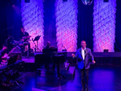A performer in a blazer stands center stage at a concert venue with a grand piano, band members, and dramatic purple and pink lighting illuminating tall architectural columns in the background.
