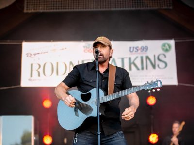 Acoustic guitarist performing on stage with professional lighting setup, including warm spotlights and stage backdrop signage visible in background.